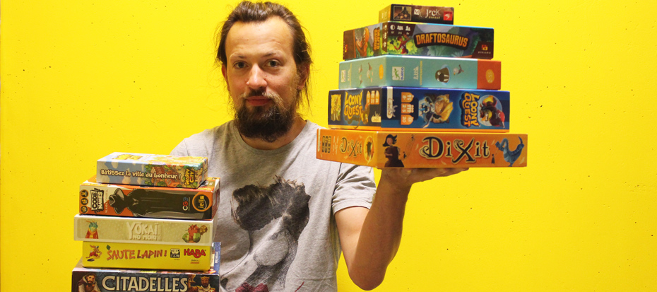 Homme portant des boites de jeux de société dans chaque main, sur fond jaune