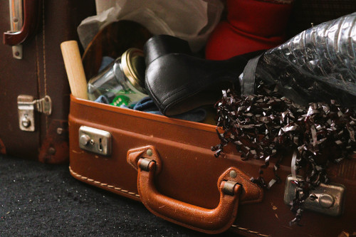 Valise en cuir marron d'où déborde des objets : chaussure à talons, boite en verre, etc.